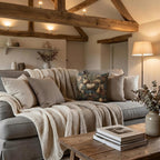 a cozy living room with a gray sofa adorned with throw pillows and a blanket, a wooden coffee table, and a lamp on a side table. The room has a rustic charm with exposed wooden beams and a vase of flowers on a shelf.